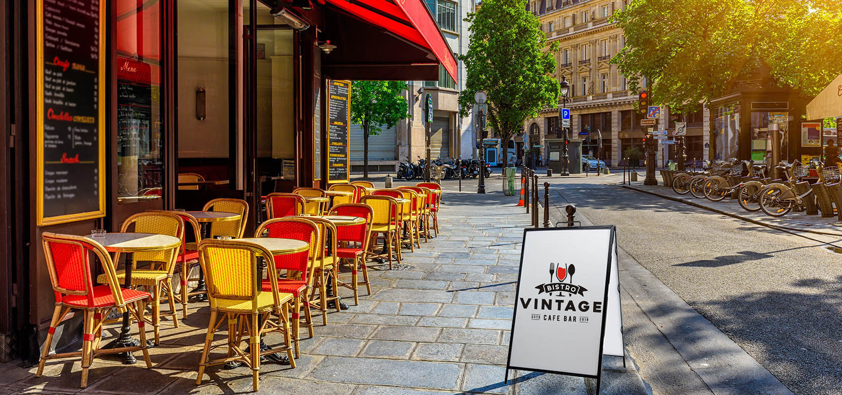 White A-frame cafe sign on a cobblestone sidewalk with outdoor restaurant seating in the background.